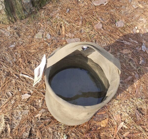 A Large Canvas Collapsible Water Bucket (ROTHCO) rests on the ground beside a tree.