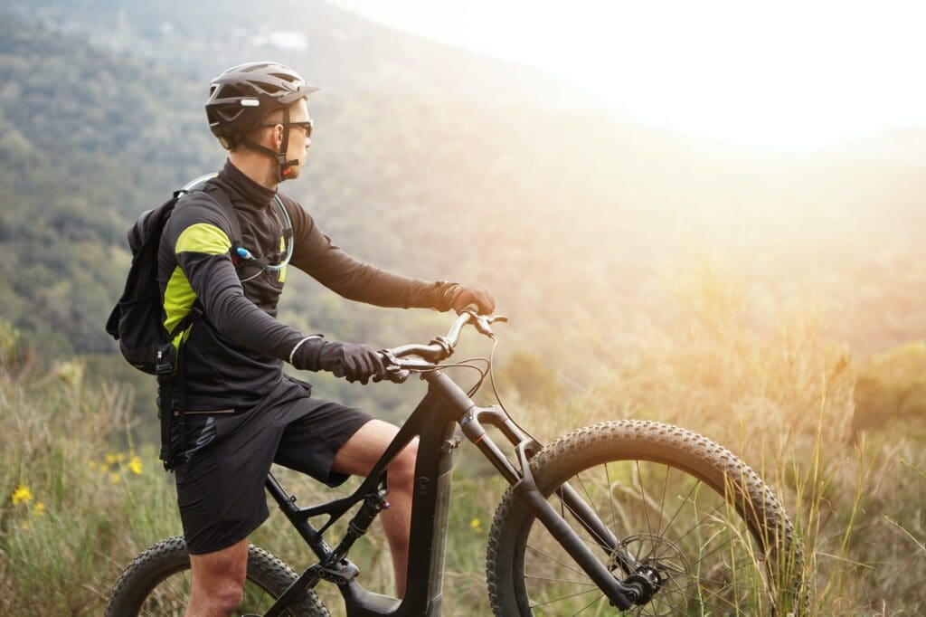 Side view of young stylish male biker in black sportswear standing on top of hill with his electric