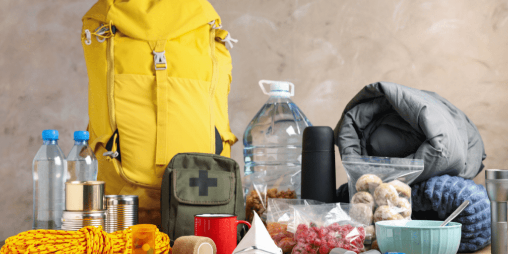 Essential items for camping or survival preparedness, including a yellow backpack, canned food, a water jug, sleeping bag, first aid kit, and other supplies with financial constraints in mind, arranged on