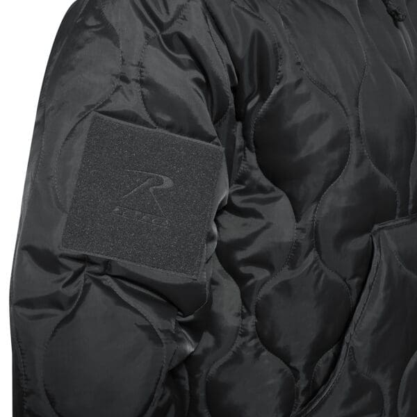 Close-up of a black quilted jacket in size 2XL, featuring a textured patch on the arm and a pocket feature. The Rothco Quilted Woobie's shiny finish and wave-like stitching patterns complement its stylish appeal.