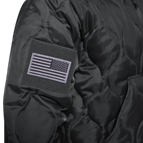 Close-up of a dark Rothco quilted jacket sleeve with a black and white American flag patch on the arm, reminiscent of their classic Woobie Hooded Sweatshirt design.