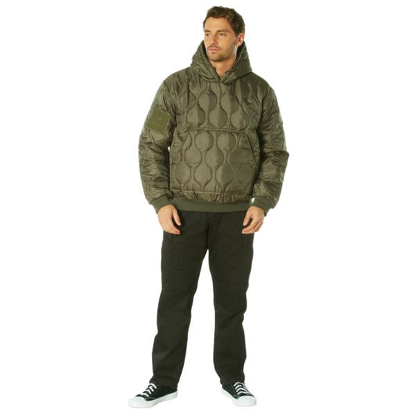 A man wearing a green quilted woobie hooded sweatshirt and black pants stands against a plain white background.