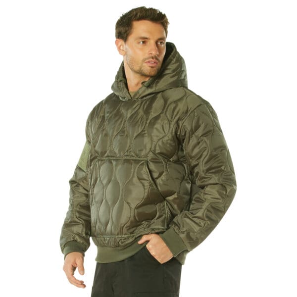 A man wearing a Rothco Quilted Woobie in olive green, featuring a front pocket, stands against a plain white background.